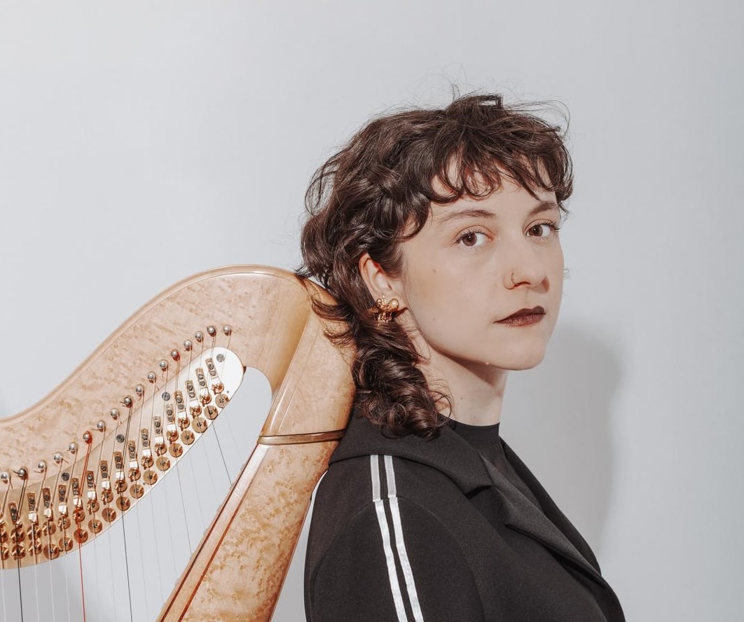 A person with short, curly hair leans against a harp, wearing a black jacket with white stripes, looking at the camera against a white background.