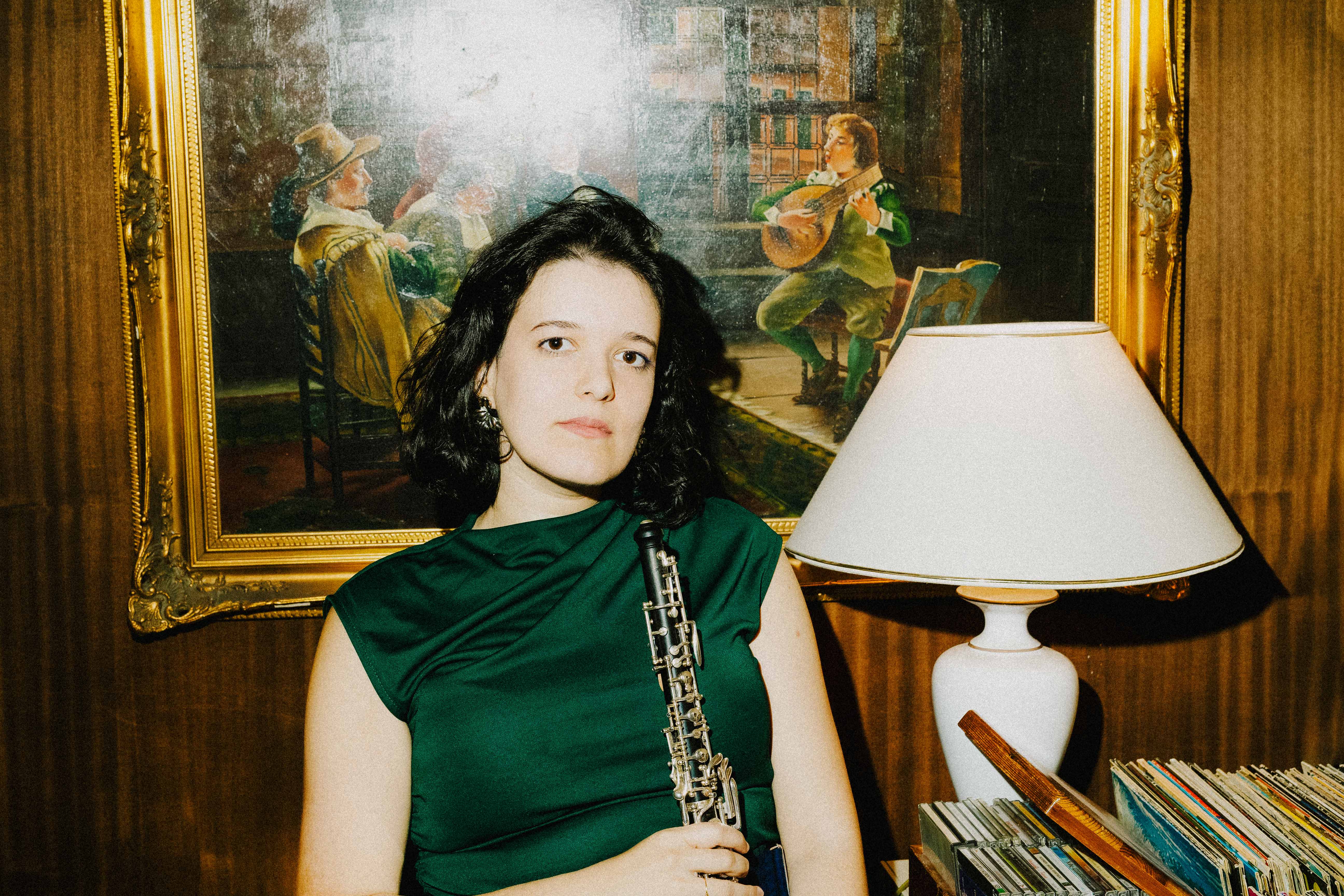 A woman in a green dress holding an oboe stands beside a lamp and framed painting in a warmly lit room.