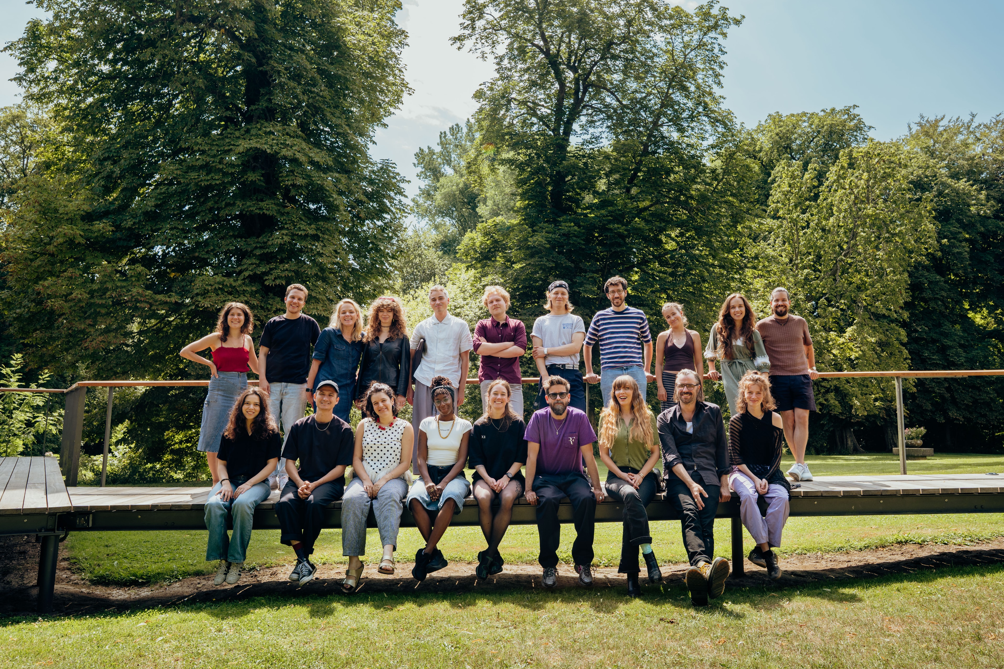 A group of 18 people posing outdoors, some sitting and some standing, with lush green trees in the background. It is a sunny day.