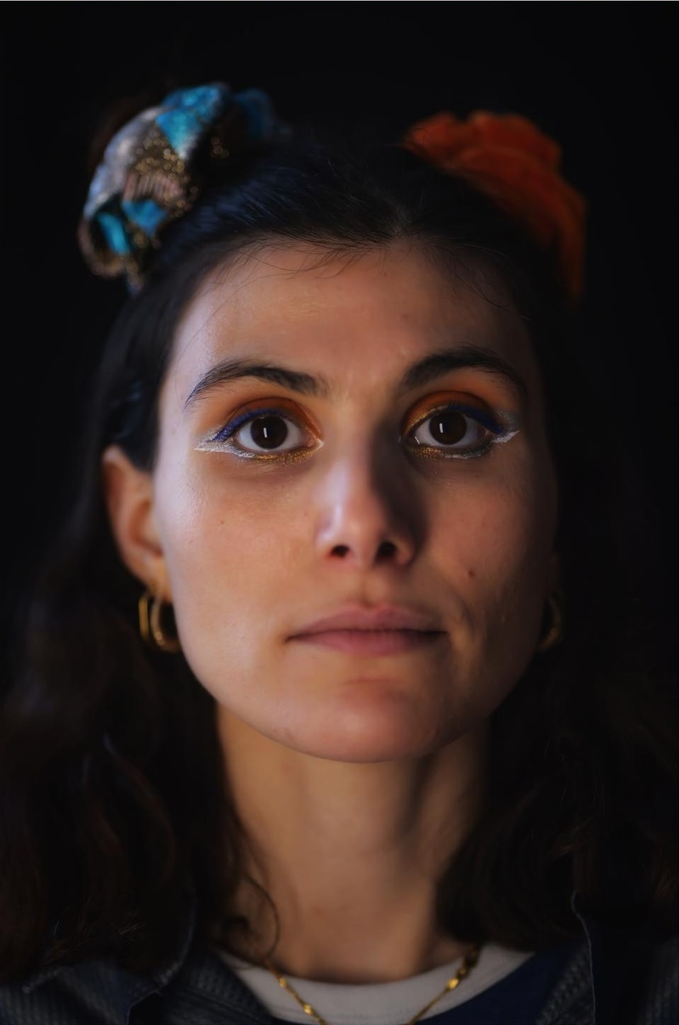 Woman with vibrant eye makeup and colorful hair accessories, gazing directly at the camera, set against a dark background.